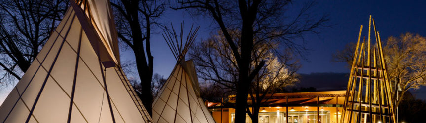 An exterior view of the Ute Indian Museum at dusk, with tepees and a wikiup sculpture in front.
